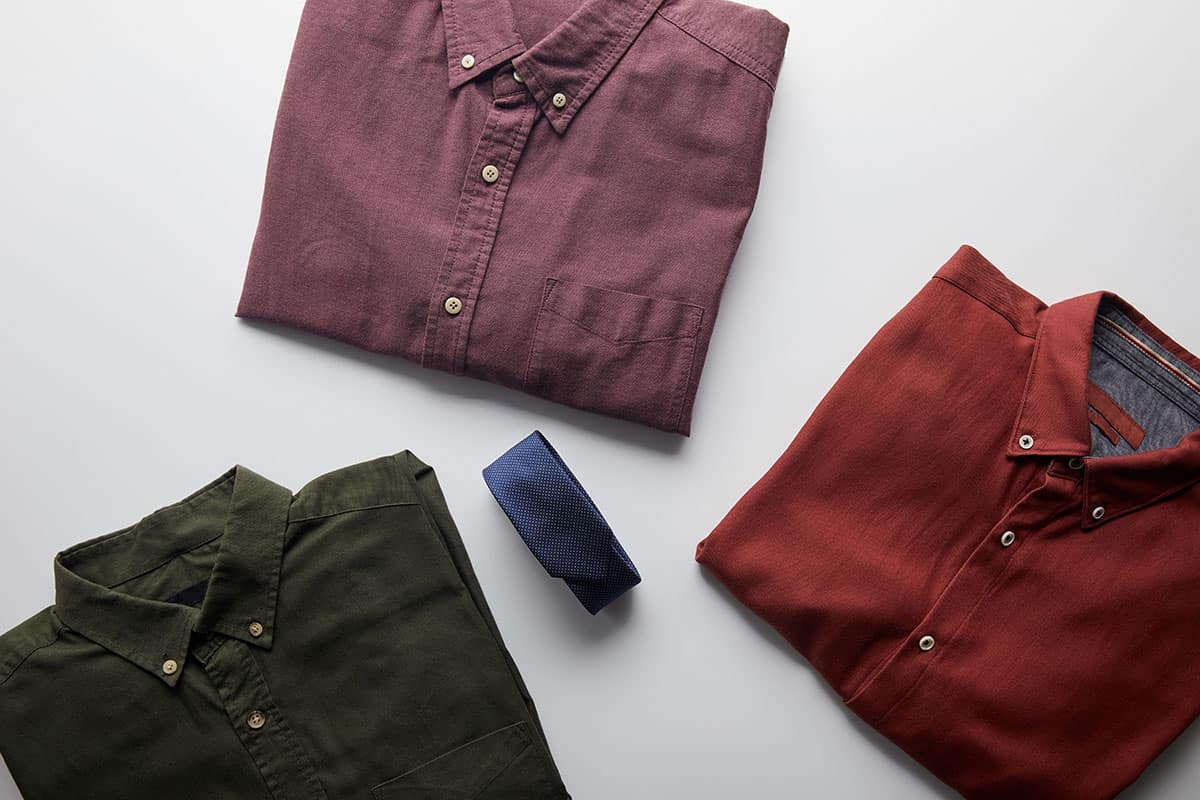 Three neatly folded San Marcos custom t-shirts in different colors: a maroon shirt, a burnt orange shirt, and an olive green shirt, accompanied by a navy blue tie, all arranged on a light gray background.